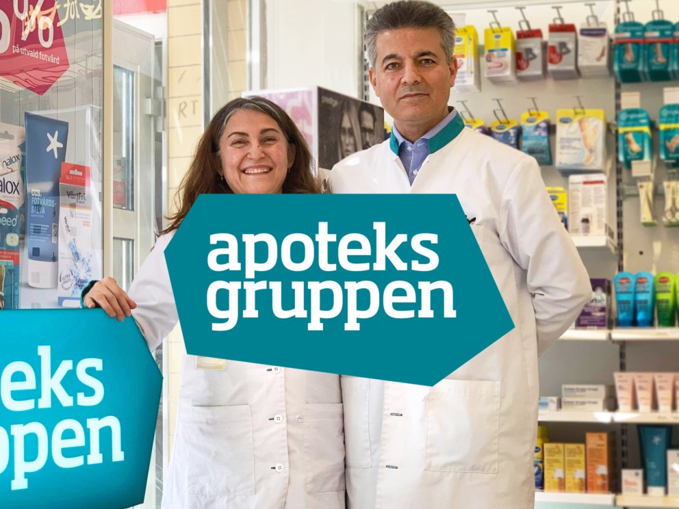 två apoteksarbetare som står bredvid varandra med apoteksgruppen logotyp i förgrunden
